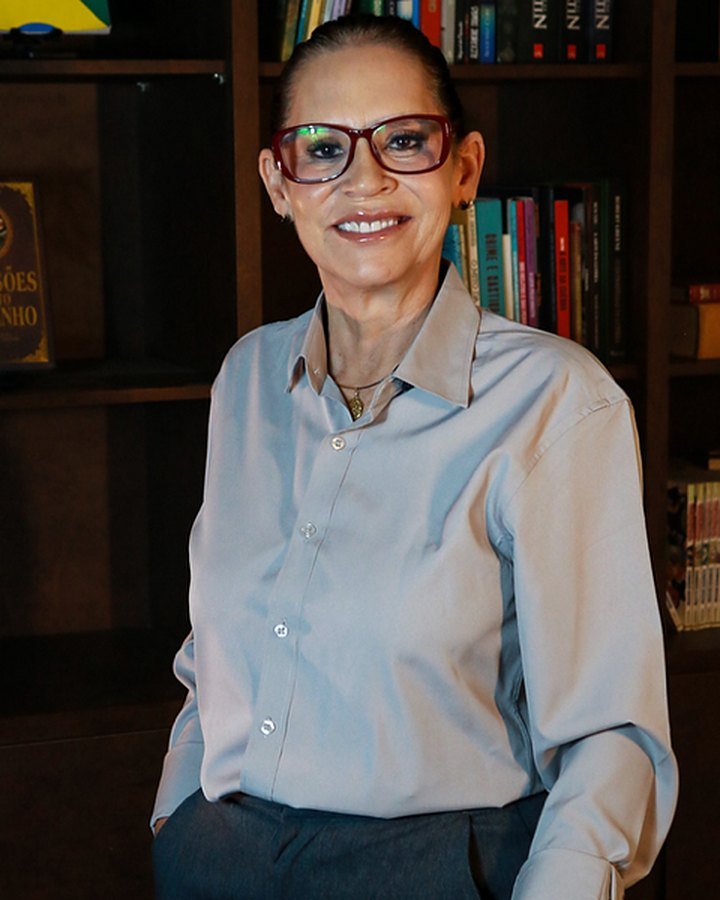 Andréia Braga em pé, de camisa clara, diante de uma estante com livros